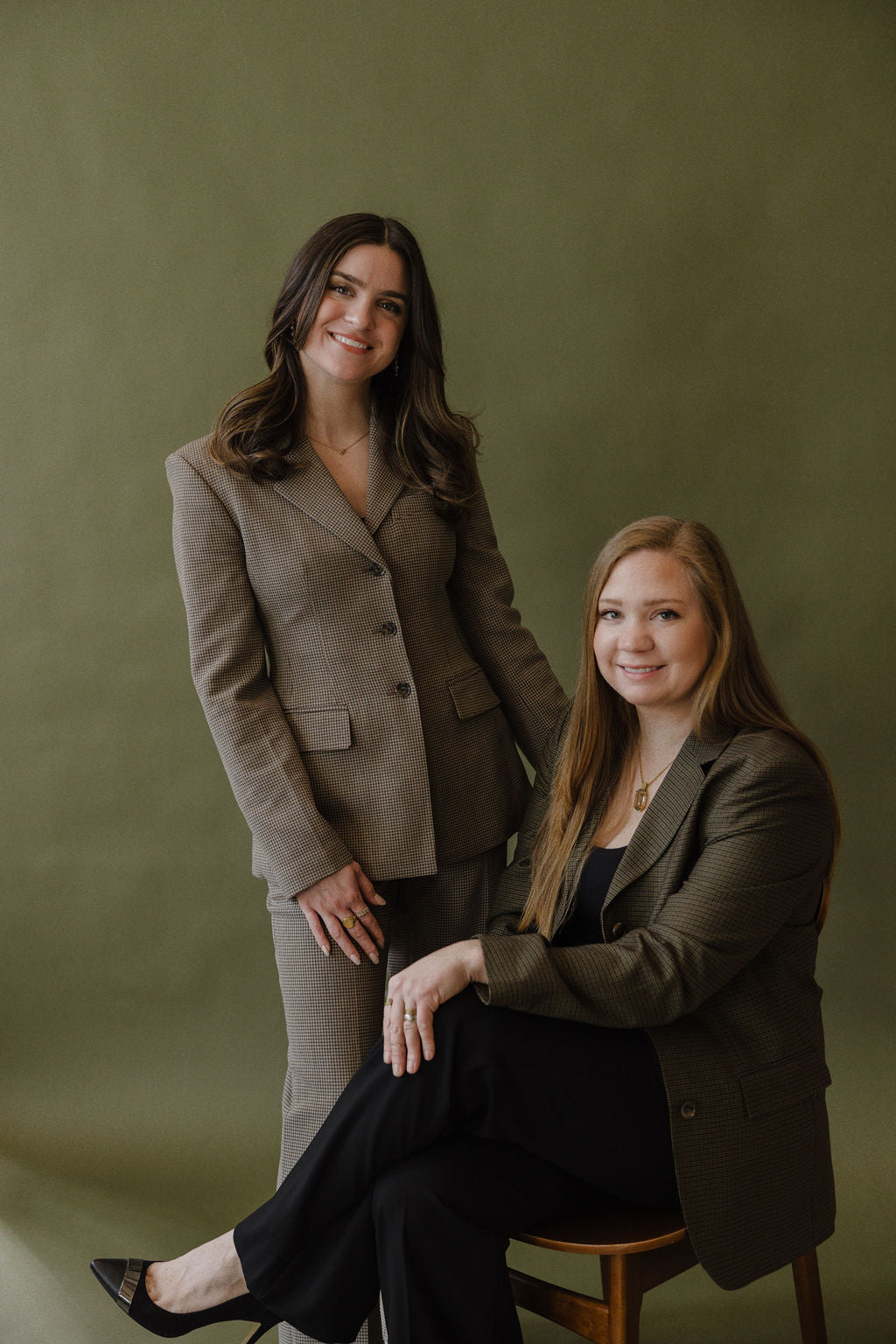 Two woman posed together in professional attire against a plain olive green background