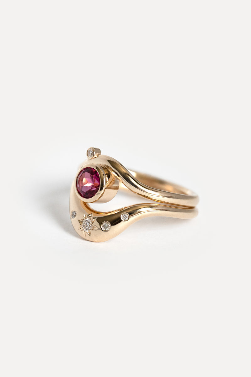 A gold ring featuring a curved band design with a central round pink gemstone and small diamond accent stones, photographed on a clean white background.