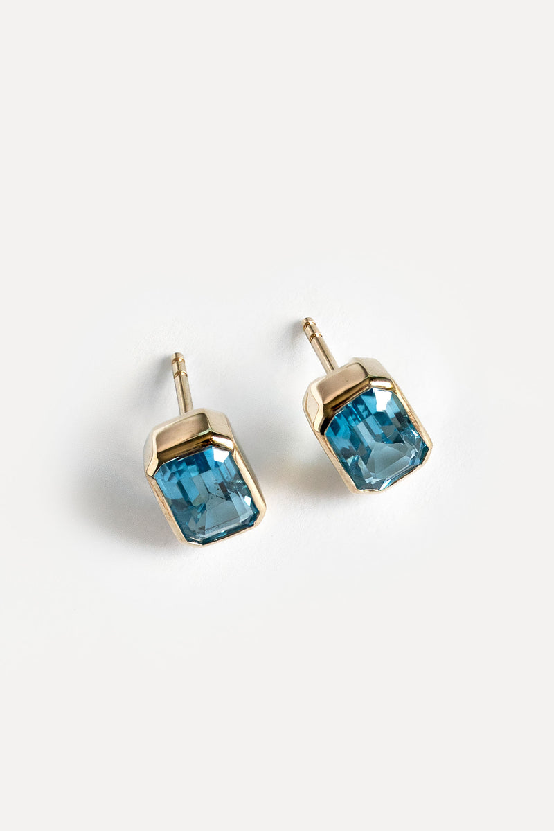A pair of gold stud earrings featuring rectangular faceted blue gemstones, displayed on a clean white background.